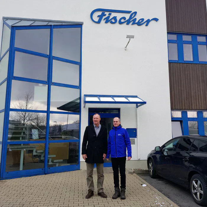 Two men standing outside a modern building with blue windows and Fischer signage, likely related to German weather instruments.