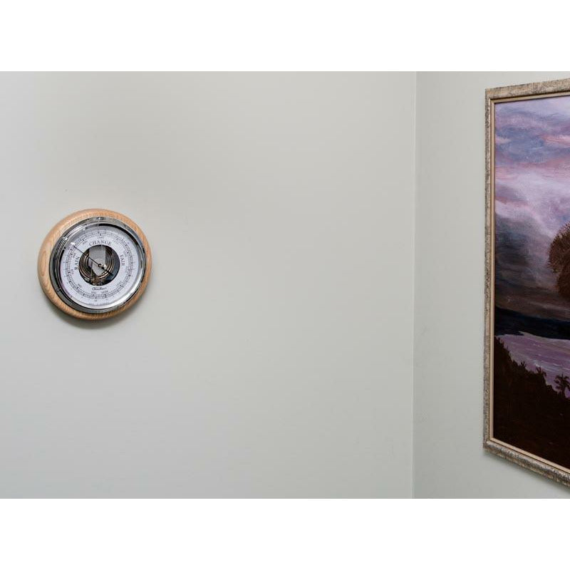 Wall-mounted German barometer with wooden frame next to a framed painting on a light wall