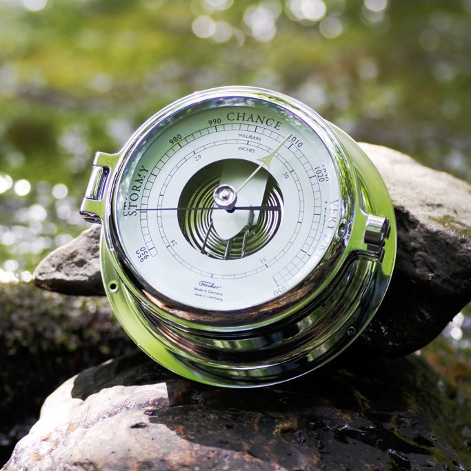 Chrome analog barometer with dial face on rocks beside water, outdoor weather instrument